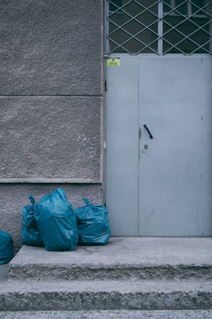 The image depicts four blue rubbish bags placed on a concrete step against the base of a building wall, which has a textured, rough grey surface. To the right, there is a closed light grey metal door